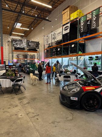 Indoor auto-parts warehouse car show with a race car hood open, tall storage racks, round tables, groups of people and a white dog.