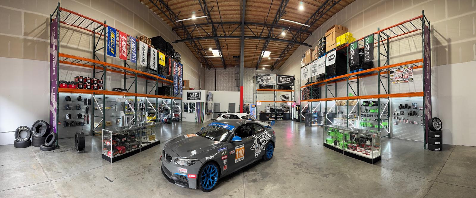 Panoramic auto parts warehouse showroom with a race‑prepped gray sports coupe (blue wheels, racing decals) centered on a polished concrete floor, surrounded by tall industrial racks, display cases of performance parts and stacked tires under bright overhead lighting.