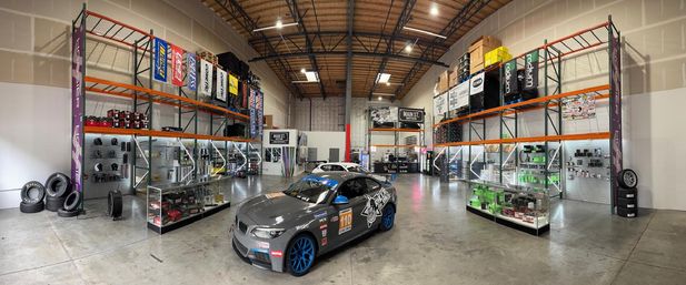 Panoramic auto parts warehouse showroom with a race‑prepped gray sports coupe (blue wheels, racing decals) centered on a polished concrete floor, surrounded by tall industrial racks, display cases of performance parts and stacked tires under bright overhead lighting.