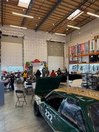 Indoor automotive warehouse hosting a holiday car show — green #222 race car with open hood in foreground, high wooden-beamed ceiling with string lights, vendor tables, people mingling and shelving with banners in the background.