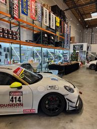 White track-ready sports car #414 inside an indoor motorsports warehouse — tall shelving with performance parts and banners, lit display racks, and a snacks/refreshments table by a small decorated tree.