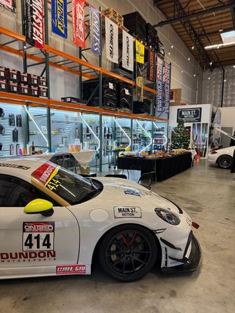 White track-ready sports car #414 inside an indoor motorsports warehouse — tall shelving with performance parts and banners, lit display racks, and a snacks/refreshments table by a small decorated tree.