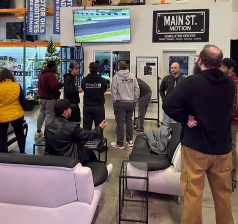 Group of people in a warehouse-style indoor racing simulator lounge watching a wall-mounted TV showing a racetrack, with sofas, leaderboard monitors, auto-supply shelves and a small Christmas tree.