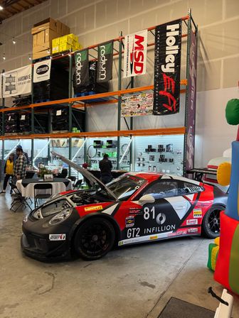 GT2 race car #81 in matte black and red with its hood open on display inside a performance-parts warehouse showroom, tall shelving with hanging banners and product walls and people browsing nearby.