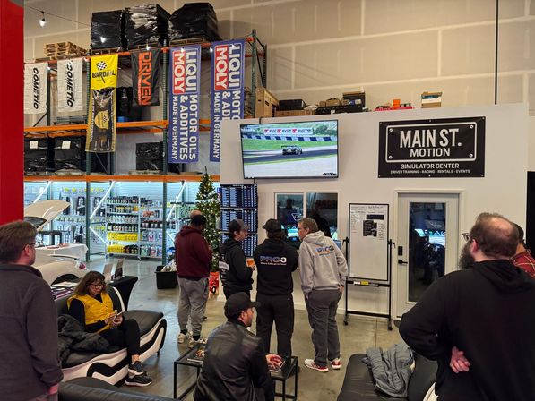 Indoor racing simulator lounge in an auto warehouse — a small crowd watches a live race on a large wall screen, with simulator rooms, shelving of automotive fluids and parts, banners and a holiday tree in the background.