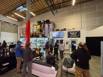 Car enthusiasts gathered in a warehouse-style automotive showroom and simulator lounge, watching a racing simulator on a wall screen amid sofas, sports cars, tall parts shelving, banners and exposed high ceilings.