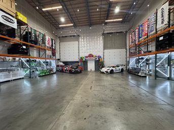 Spacious industrial garage warehouse interior with concrete floor and high ceiling, shelving and illuminated display racks along the sides, two race cars parked near large roll-up doors and a colorful inflatable arch at the back.