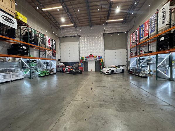 Spacious industrial garage warehouse interior with concrete floor and high ceiling, shelving and illuminated display racks along the sides, two race cars parked near large roll-up doors and a colorful inflatable arch at the back.