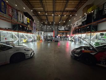 Warehouse-style automotive showroom with high ceilings and string lights, two race-style sports cars parked left and right, illuminated parts shelves lining the walls, concrete floor and a small decorated Christmas tree near center.