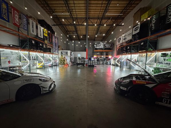 Warehouse-style automotive showroom with high ceilings and string lights, two race-style sports cars parked left and right, illuminated parts shelves lining the walls, concrete floor and a small decorated Christmas tree near center.