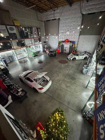 High-angle view of an industrial warehouse showroom with three sports cars, string lights, round standing tables, a lit Christmas tree and inflatable holiday backdrop — festive auto shop event space.