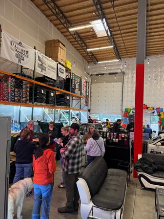 Busy warehouse-style showroom with tall metal shelving and stacked boxes, shoppers chatting near product displays and couches, string lights and a large garage door, and a dog on a leash.