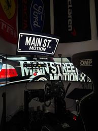 Home sim-racing cockpit with curved triple monitors showing 'MAIN ST. MOTION' graphics, force-feedback wheel, pedal set, shifter and an OMP racing seat under automotive flags — immersive gaming rig.