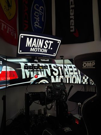 Home sim-racing cockpit with curved triple monitors showing 'MAIN ST. MOTION' graphics, force-feedback wheel, pedal set, shifter and an OMP racing seat under automotive flags — immersive gaming rig.