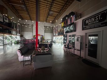 Warehouse-style indoor racing simulator lounge with high ceilings, string lights, cozy seating and a white sports car on display among neon-lit gear racks.