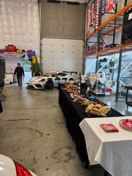 Festive warehouse garage scene with a white race car by a 'Merry Christmas' inflatable arch and a long catered buffet table with sandwiches, fruit, shelving and a large overhead door.