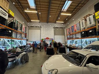 White sports car in foreground inside a high‑ceiling auto shop warehouse set up for an indoor car meet — string lights, parts shelving and banners, people mingling around cocktail tables and food stations.