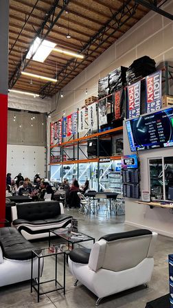 Automotive warehouse event space with high industrial ceiling, racing product banners on tall racks, black-and-white lounge sofas, folding tables of people, and a large digital timing screen.