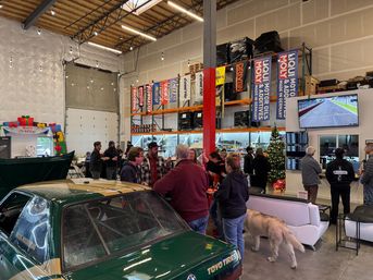 Warehouse-style auto shop holiday car meet with a green race car in the foreground, people mingling near parts shelving and banners, a decorated Christmas tree, TV showing a racetrack, and a dog walking through the space.