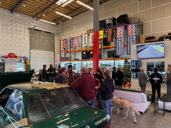 Warehouse-style auto shop holiday car meet with a green race car in the foreground, people mingling near parts shelving and banners, a decorated Christmas tree, TV showing a racetrack, and a dog walking through the space.