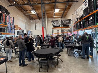 Bustling indoor industrial warehouse event with attendees mingling around black-clothed round tables and folding chairs, tall stocked metal shelves, exposed wood ceiling beams and string lights overhead.