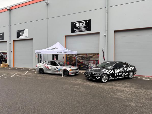 Two modified BMW sedans parked outside a gray industrial auto garage with roll-up bays—silver race car #36 under a white pop-up tent and a black checkered promotional car on a paved lot.