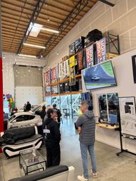 High-ceiling auto shop warehouse with open service bays and cars, people chatting by couches, shelving stacked with racing banners and pallets, and a large wall TV showing a car on track.