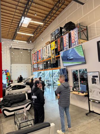 High-ceiling auto shop warehouse with open service bays and cars, people chatting by couches, shelving stacked with racing banners and pallets, and a large wall TV showing a car on track.