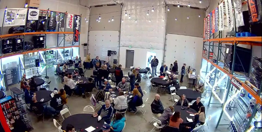 Wide-angle view of an indoor warehouse event with round tables, folding chairs, string lights, product racks and groups of people mingling