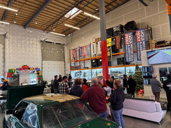Holiday-themed indoor auto shop meet with people gathered around a green race car in a high-ceiling garage, shelving with auto parts and banners, tools displays and a decorated Christmas tree.