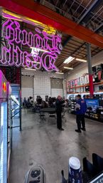 Neon pink overhead sign lighting an industrial warehouse showroom where folding tables and chairs host a community event, people mingling by auto‑parts shelves and banners under high skylights.