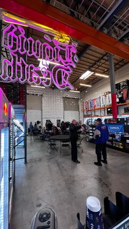 Neon pink overhead sign lighting an industrial warehouse showroom where folding tables and chairs host a community event, people mingling by auto‑parts shelves and banners under high skylights.