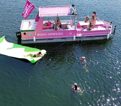 Pink pontoon party boat anchored on a tree-lined lake with people sunbathing and socializing on deck, swimmers nearby and someone lounging on a green inflatable ramp on a sunny summer day.