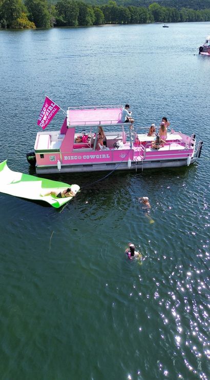 Pink pontoon party boat anchored on a tree-lined lake with people sunbathing and socializing on deck, swimmers nearby and someone lounging on a green inflatable ramp on a sunny summer day.