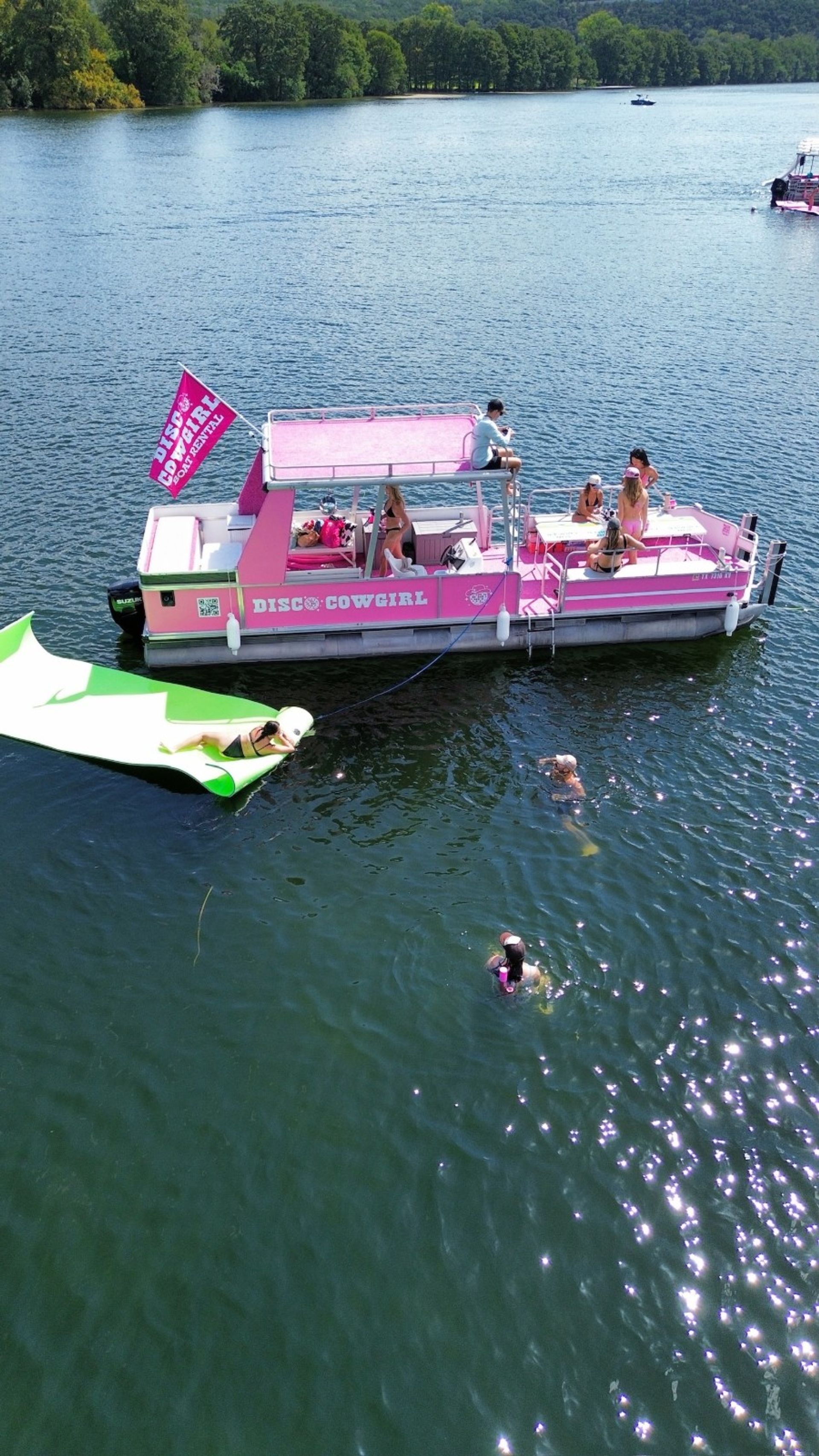 Pink pontoon party boat anchored on a tree-lined lake with people sunbathing and socializing on deck, swimmers nearby and someone lounging on a green inflatable ramp on a sunny summer day.