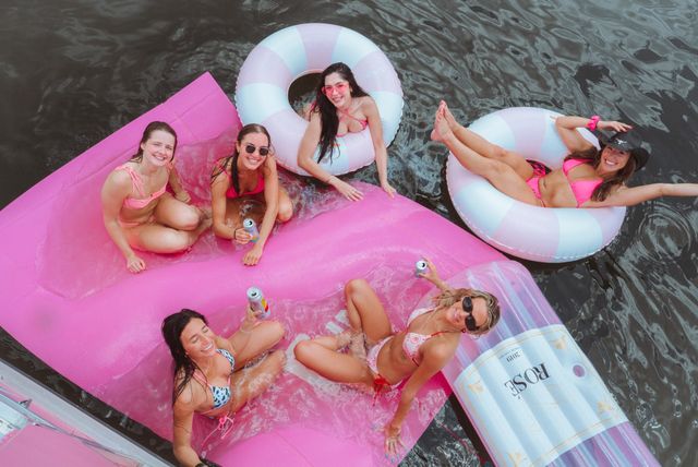 Group of friends in bright bikinis relaxing on pink inflatable floats and pastel donut tubes in a lake, smiling and sipping canned drinks on a sunny summer day.