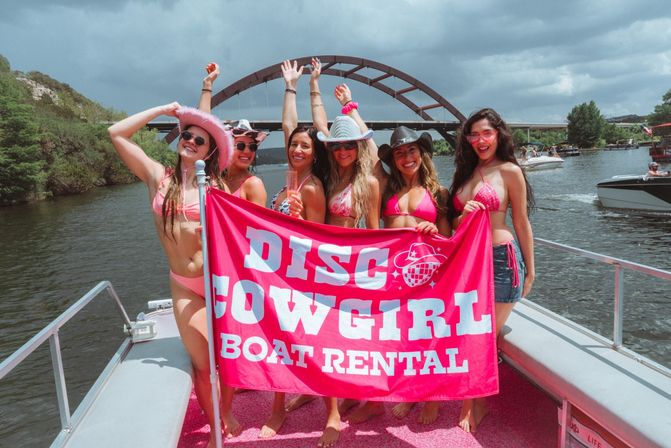 Six women in pink bikinis and cowgirl hats posing on a pink-deck rental boat, holding a bright pink banner advertising a boat rental, smiling in front of an arched bridge on a sunny river with other boats nearby.