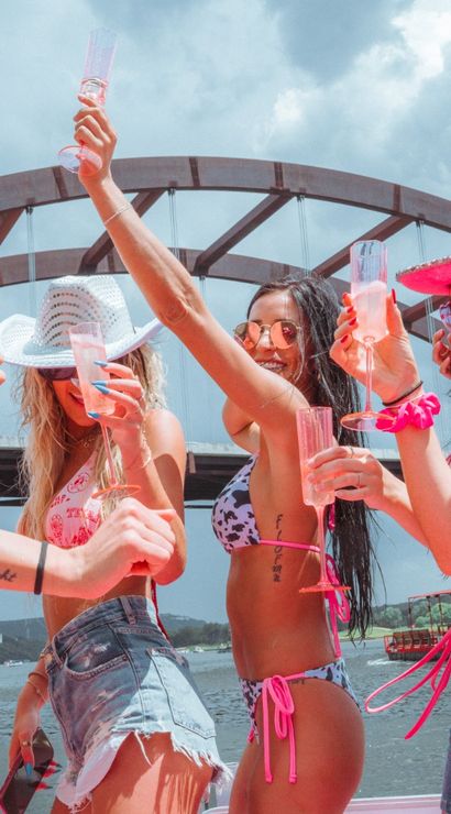 Group of women in pink bikinis and cowboy hats raising champagne flutes on a boat beneath a large arched bridge over a lake, lively summer boat party vibe