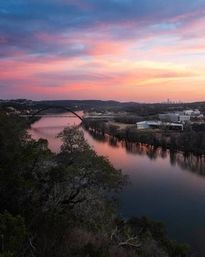 Sunset-painted pink and blue sky over a calm river with an arch bridge and distant city skyline, reflections on the water and tree-lined banks seen from a rocky overlook.