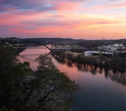 Sunset-painted pink and blue sky over a calm river with an arch bridge and distant city skyline, reflections on the water and tree-lined banks seen from a rocky overlook.