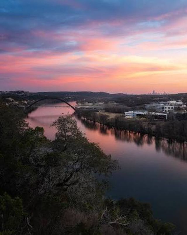 Sunset-painted pink and blue sky over a calm river with an arch bridge and distant city skyline, reflections on the water and tree-lined banks seen from a rocky overlook.