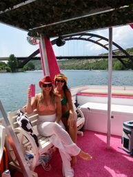 Two friends in swimsuits and cowboy hats relaxing on a pink-decorated pontoon boat, holding a drink on a sunny river beneath an arched bridge with green shoreline