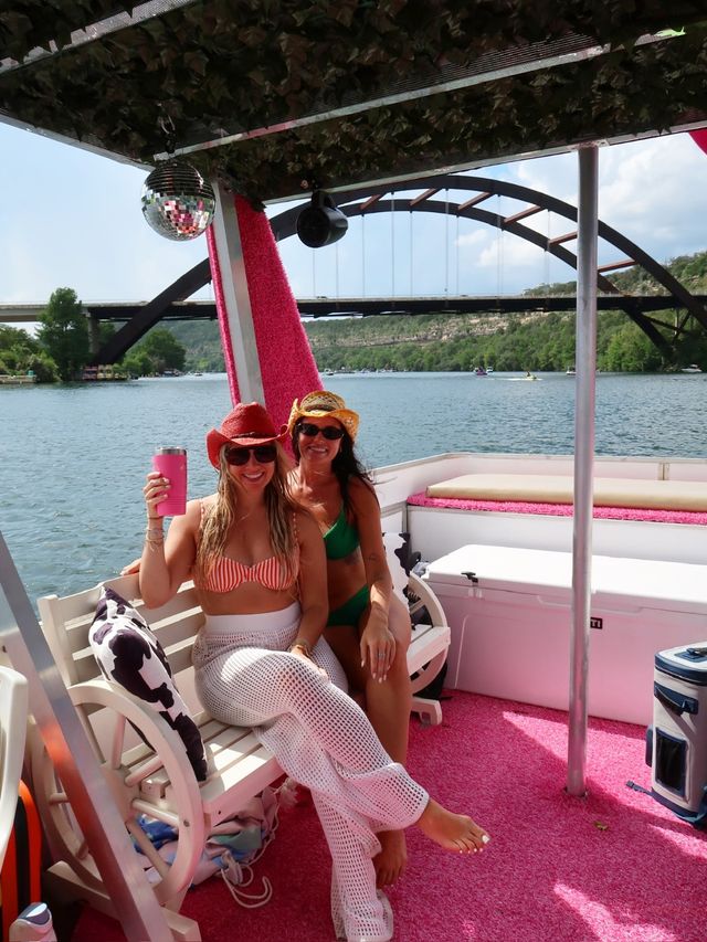 Two friends in swimsuits and cowboy hats relaxing on a pink-decorated pontoon boat, holding a drink on a sunny river beneath an arched bridge with green shoreline