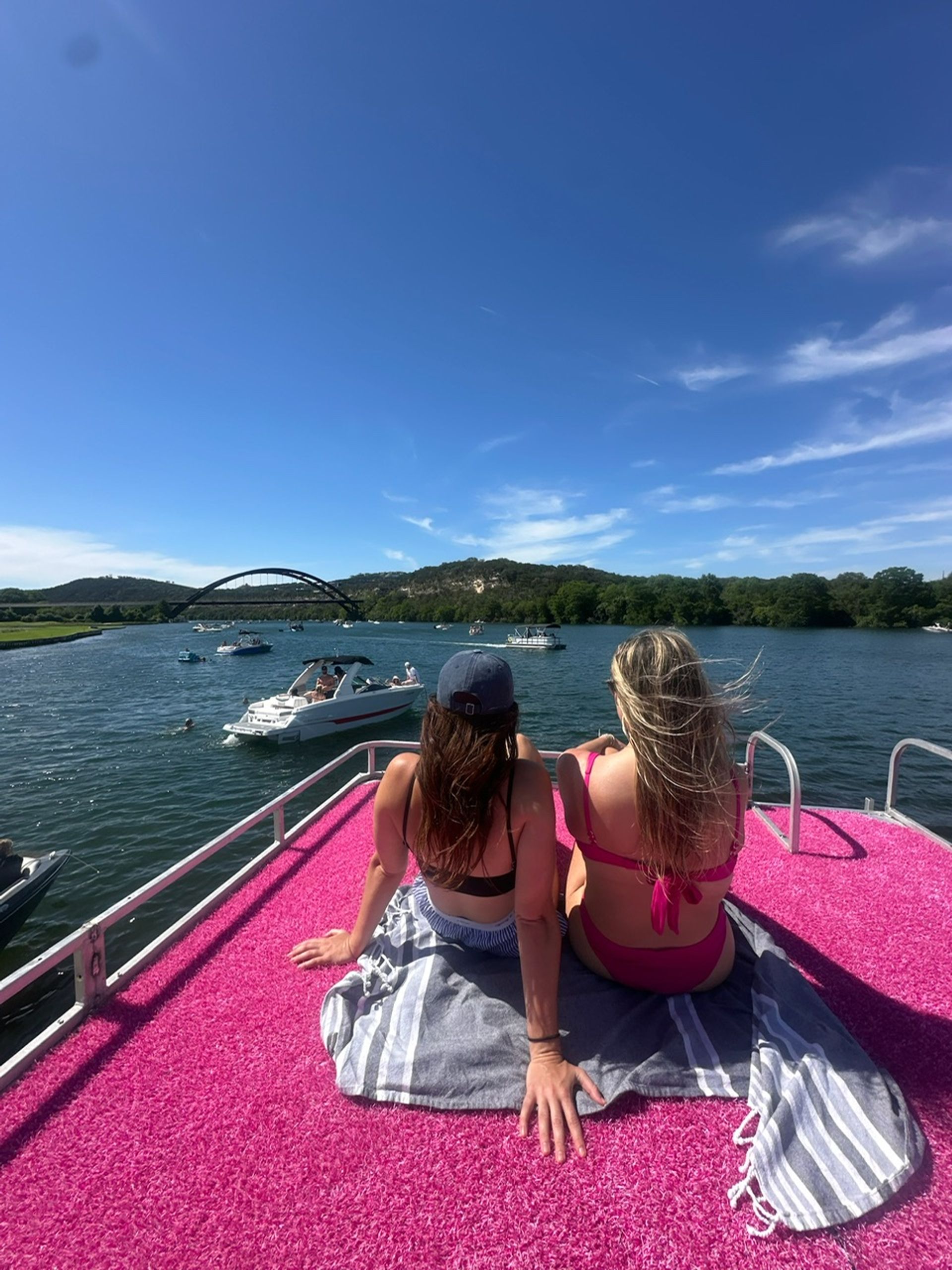Two friends relaxing on a bright pink boat deck, facing a scenic lake dotted with boats, an arched bridge and tree-covered hills under a clear blue summer sky.