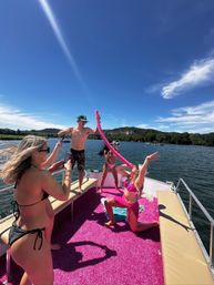 Friends partying on a pontoon boat at a sunny lake — bikinis and swim trunks, bright pink deck carpet and pool noodle, posing and taking photos with wooded shoreline.