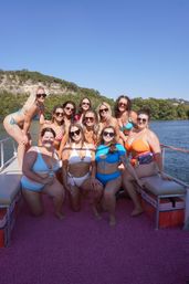 A dozen women in colorful swimsuits posing and smiling on a pontoon boat deck on a sunny lake, with tree-covered cliffs and a clear blue sky in the background.