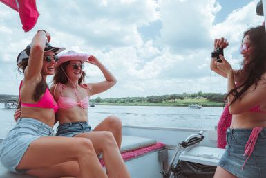Three friends on a lake boat during a sunny summer day, wearing pink bikinis, denim shorts, cowboy hats and sunglasses as one snaps a photo with a tree-lined shoreline and blue sky behind them.