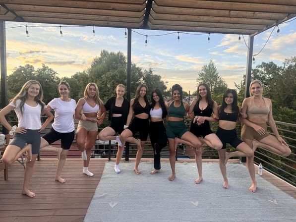 Rooftop yoga group at sunset — women in activewear holding tree pose on a wooden deck under string lights with trees and a soft evening sky in the background.