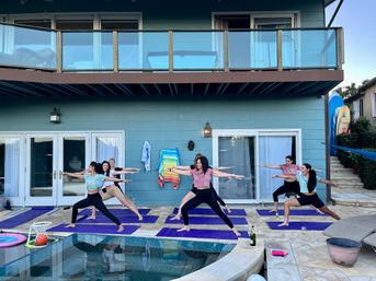 Group of women practicing poolside yoga in Warrior II on purple mats at a modern blue two-story house with balcony on a backyard patio