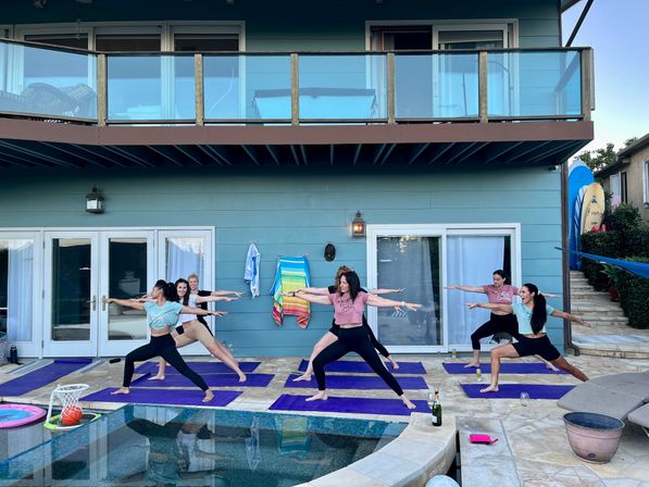 Group of women practicing poolside yoga in Warrior II on purple mats at a modern blue two-story house with balcony on a backyard patio